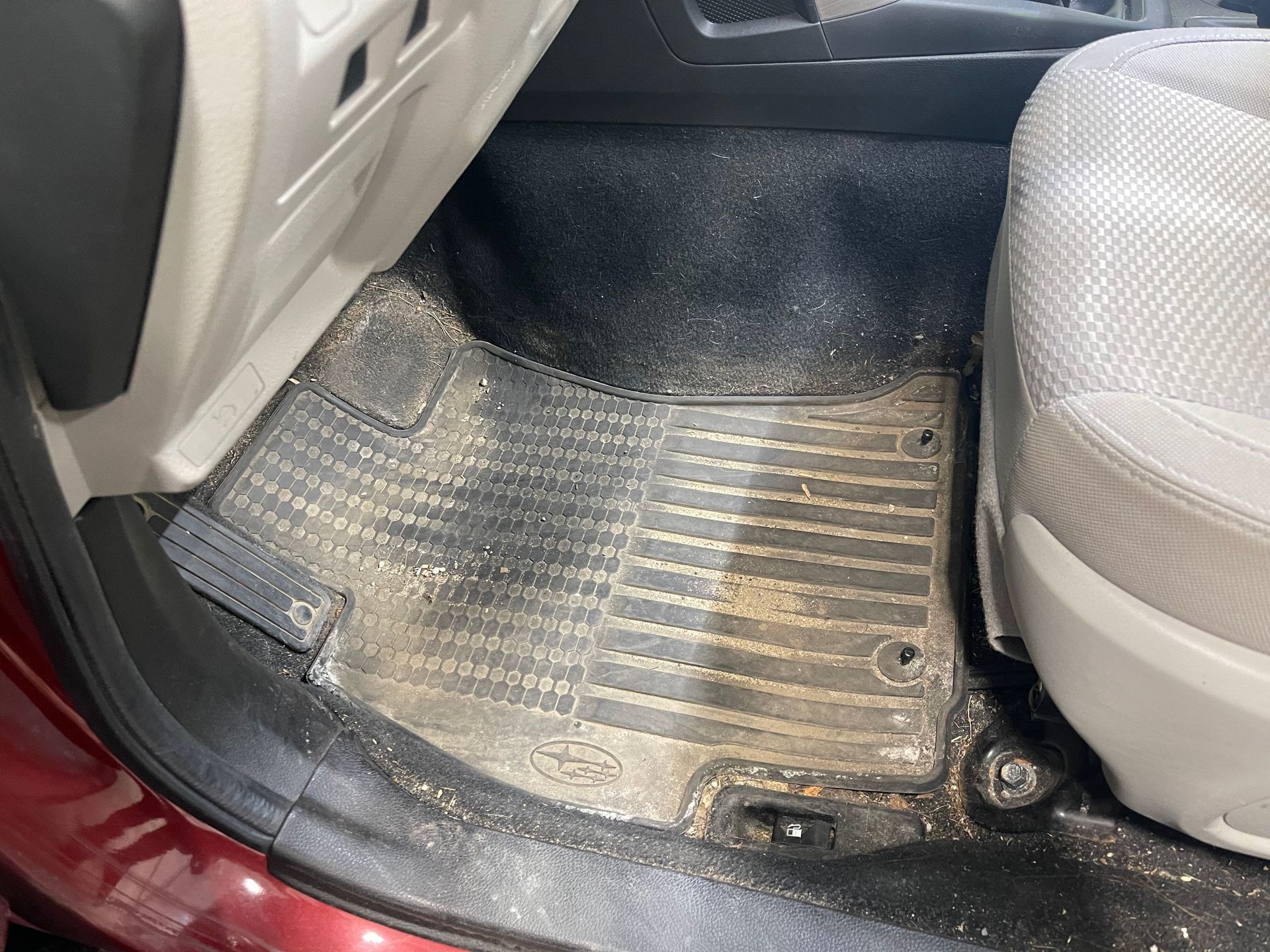 A dirty, light-colored rubber car floor mat covered in dust and debris, situated on the vehicle floor next to a seat.