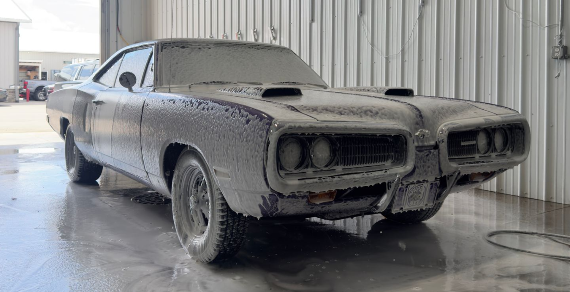 A classic muscle car covered in thick white soap suds, parked inside a garage stall with a concrete floor.