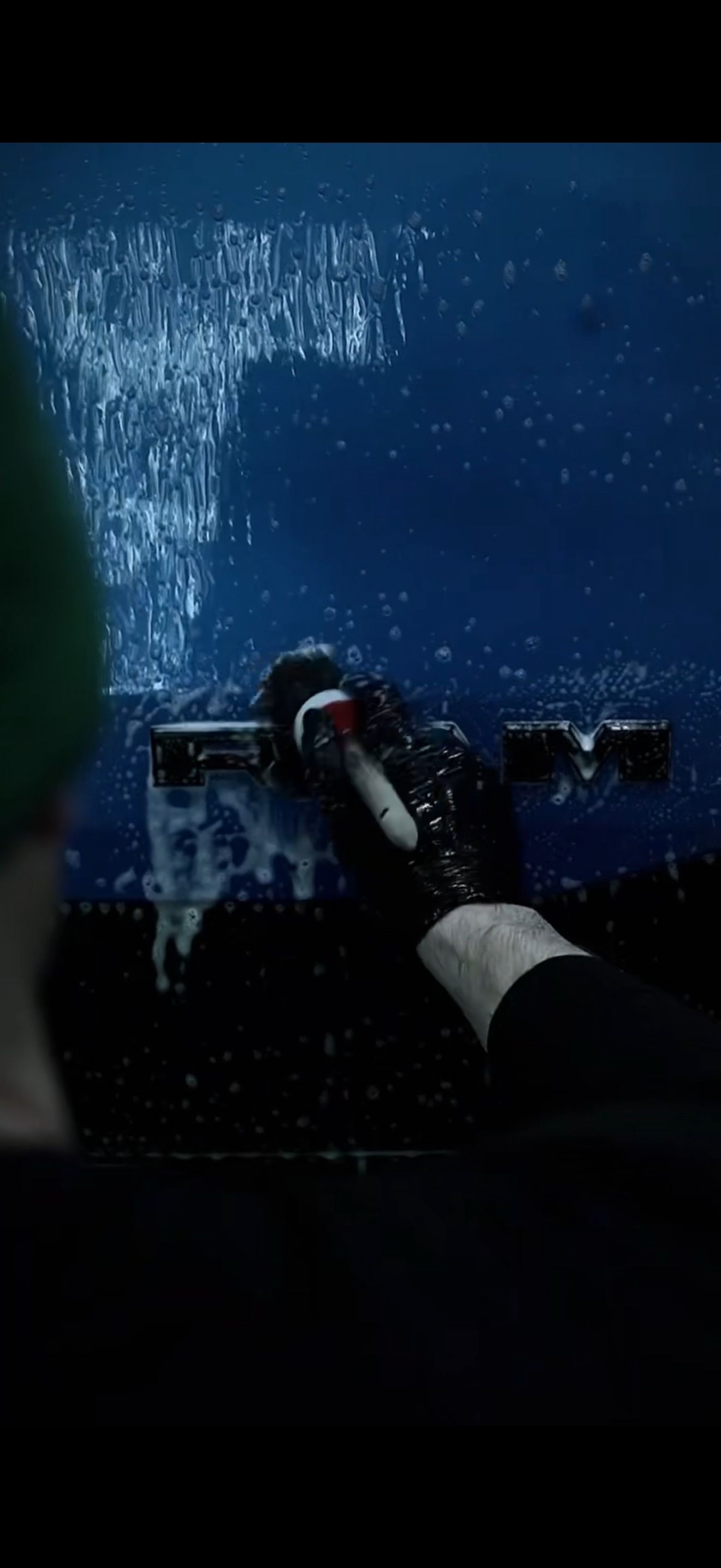 A hand wearing a black glove uses a squeegee to clean soap suds off a dark blue, glass-like surface.