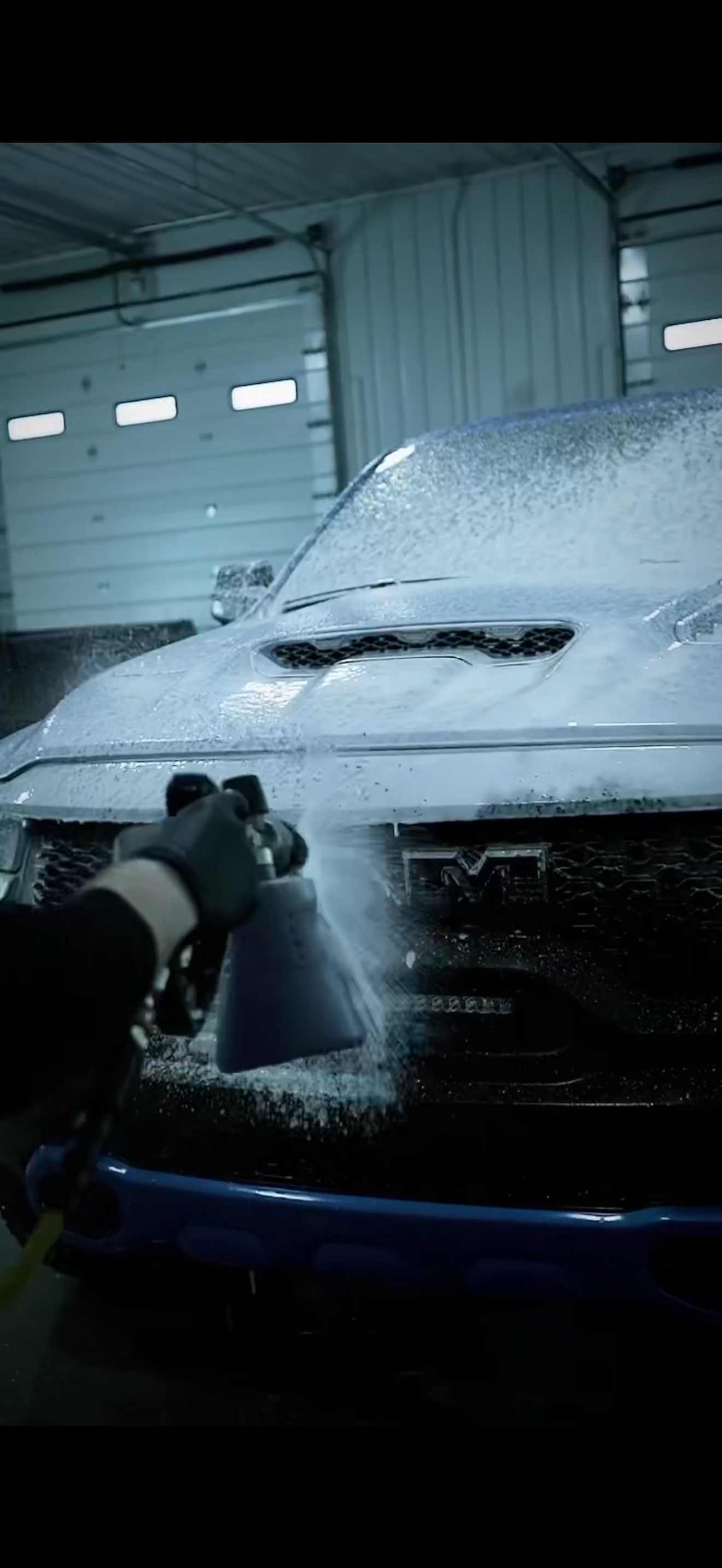 A worker uses a high-pressure hose to wash a bright blue SUV in a commercial car wash bay.