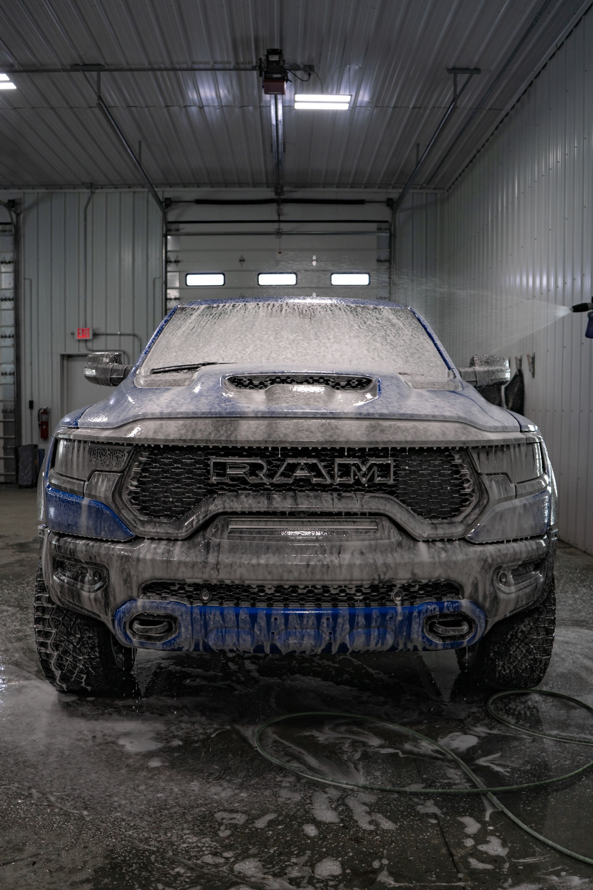 A blue RAM truck covered in thick soap suds in an indoor garage, being washed.