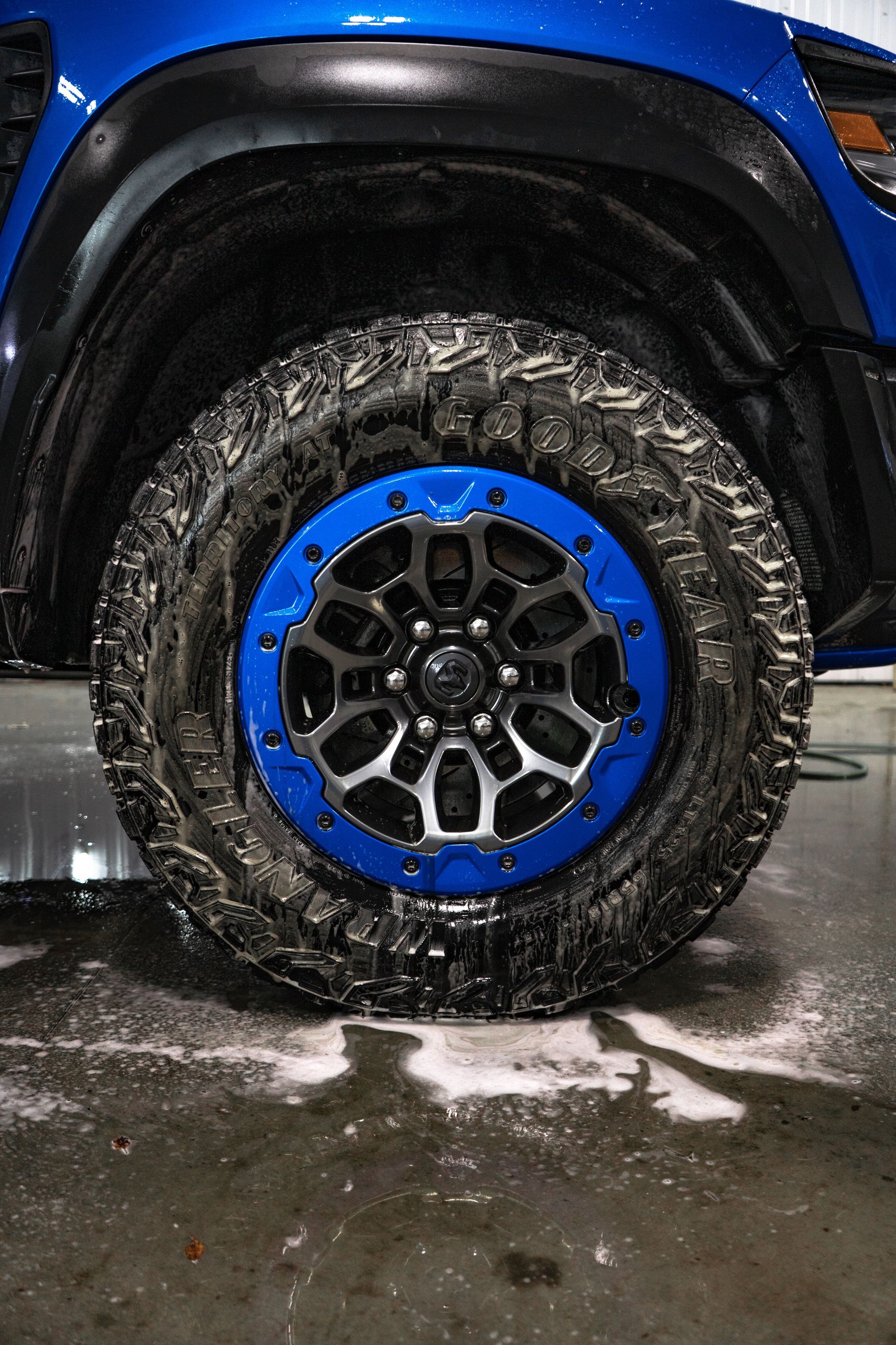 A blue truck wheel with a black and silver patterned rim, partially covered in dirt, sitting on a wet concrete floor.