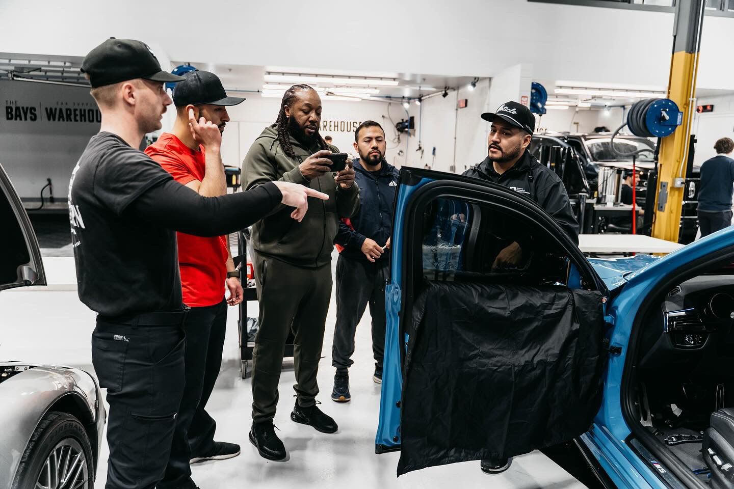 Four people stand in an auto workshop, observing and discussing work on the open door of a blue vehicle.