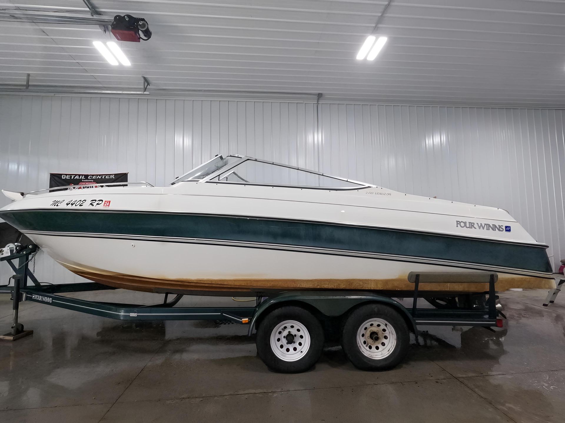 A white and dark green motorboat on a dual-axle trailer inside a warehouse with bright overhead lighting.