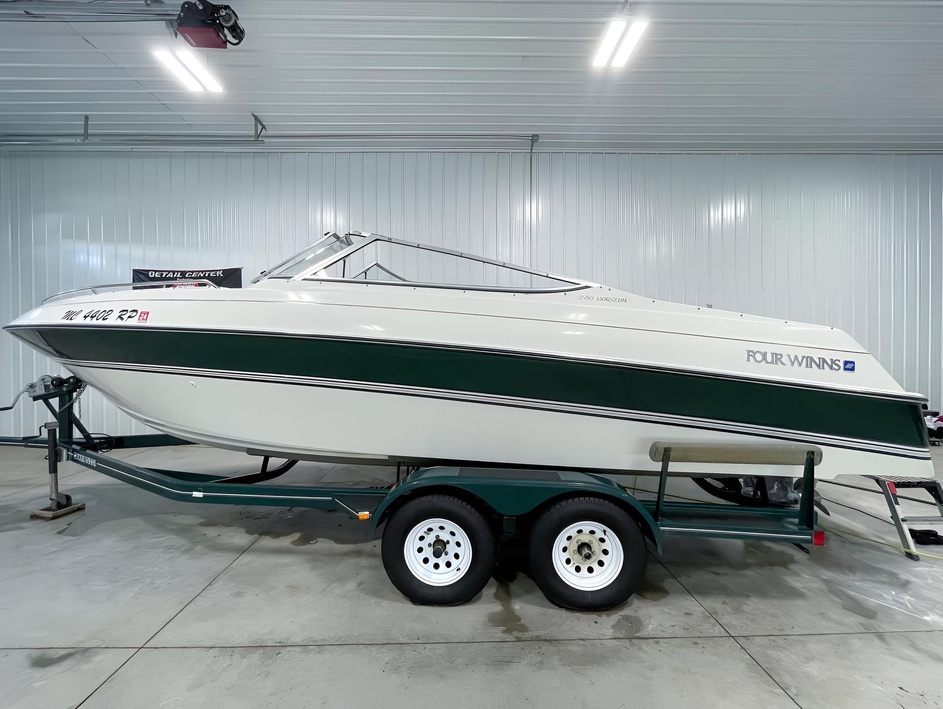 A white and dark green recreational boat on a dual-axle trailer, parked inside a large, industrial-style garage.