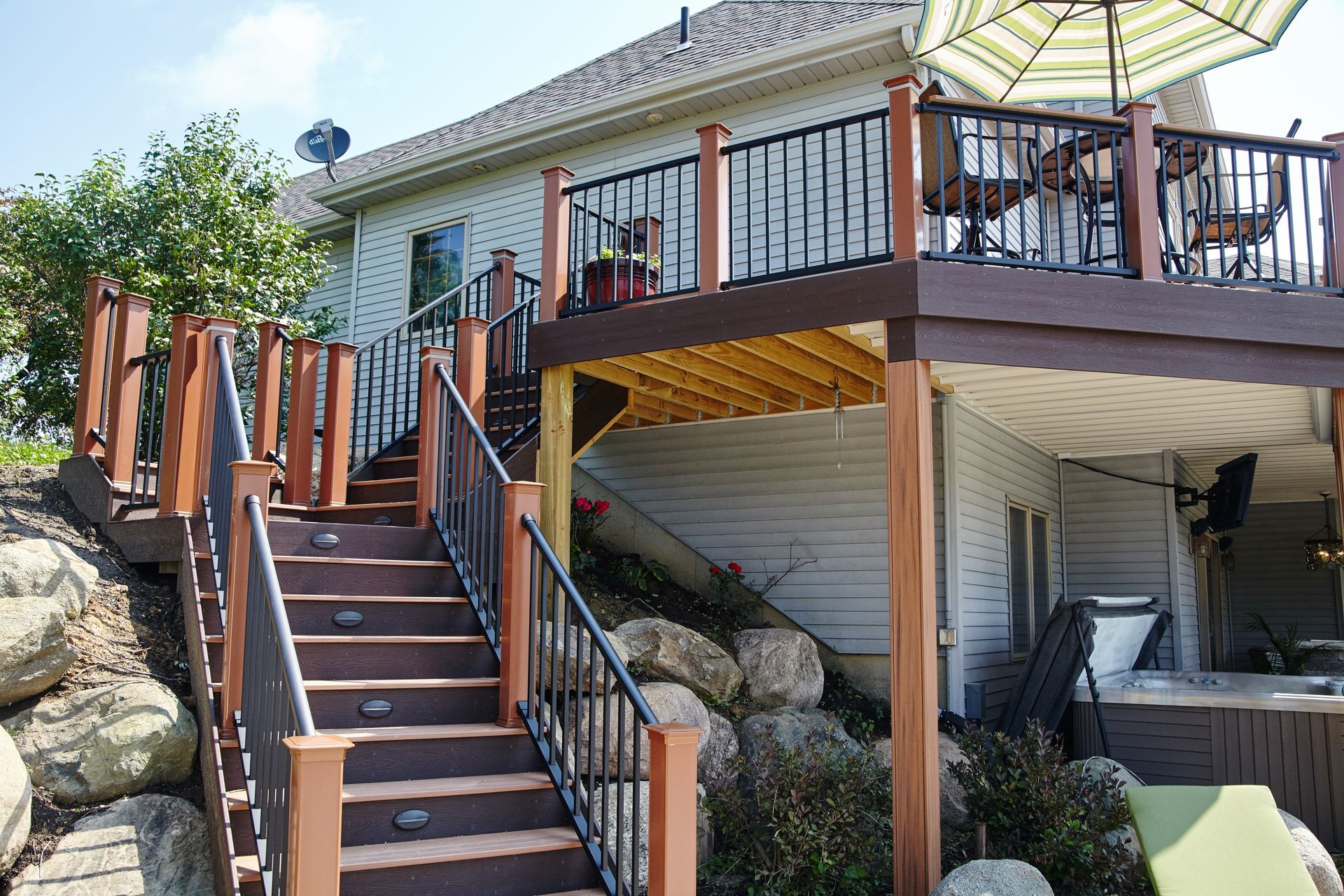 Wooden outdoor stairs leading to a deck with patio furniture beside a white house, surrounded by rocks and trees