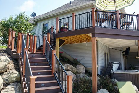 Wooden outdoor stairs leading to a deck with patio furniture beside a white house, surrounded by rocks and trees
