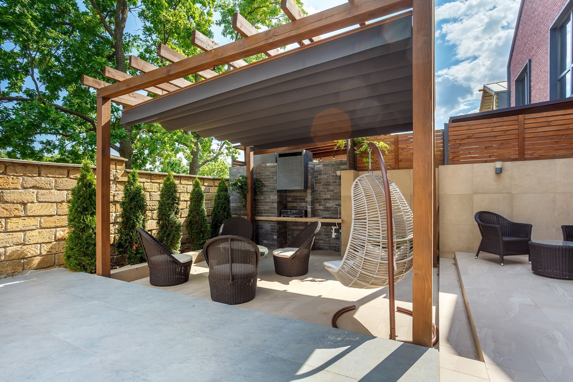 Outdoor patio with wooden pergola, stone wall, wicker seating, and potted plants under a bright sky