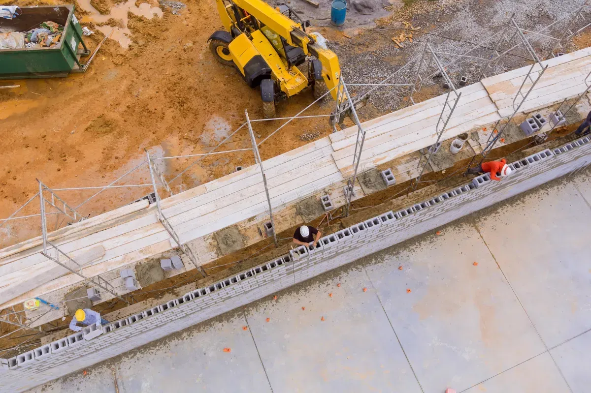 Construction site with yellow excavator beside a concrete wall and workers on a paved area.
