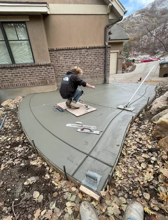 Worker smoothing fresh concrete sidewalk beside a house, with tools and forms visible.