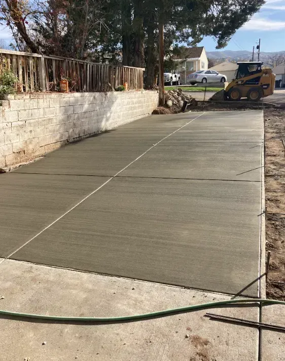 Freshly poured concrete driveway beside a stone wall, with a hose across the foreground and a skid steer at the end.