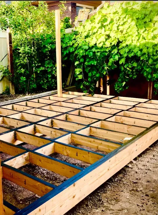 Wooden deck frame under construction in a backyard garden, with grid joists and green foliage nearby