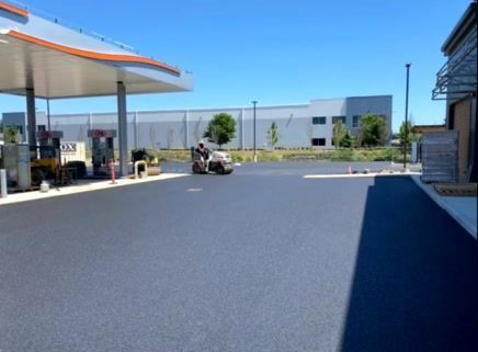 Asphalt pavement being rolled at a gas station under a blue sky, with a building in the background.