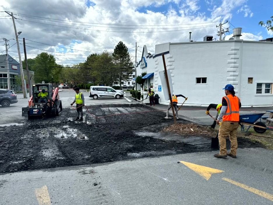 Road work: workers in vests repair asphalt; a white building and vehicles in background.