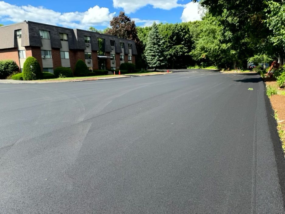 Newly paved asphalt parking lot in front of a multi-story brick apartment building on a sunny day.