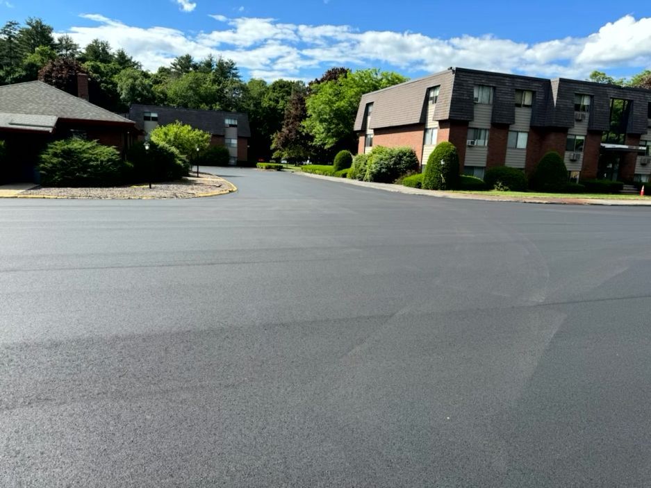 Newly paved asphalt road in front of brick apartment buildings and lush trees under a blue sky.