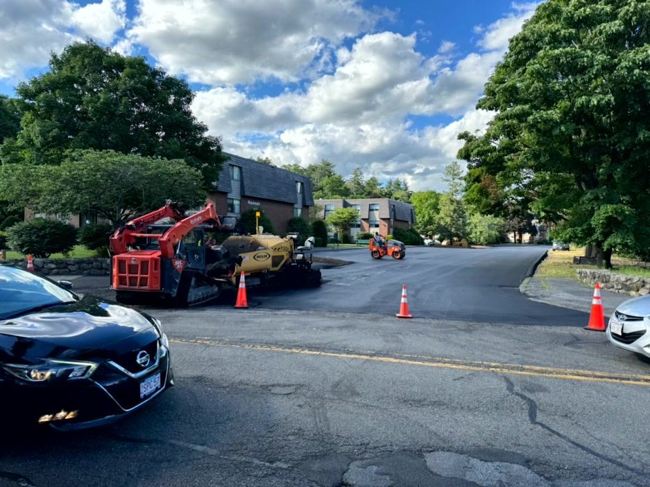 Construction equipment on a street; orange cones, parked cars, residential area, cloudy sky.