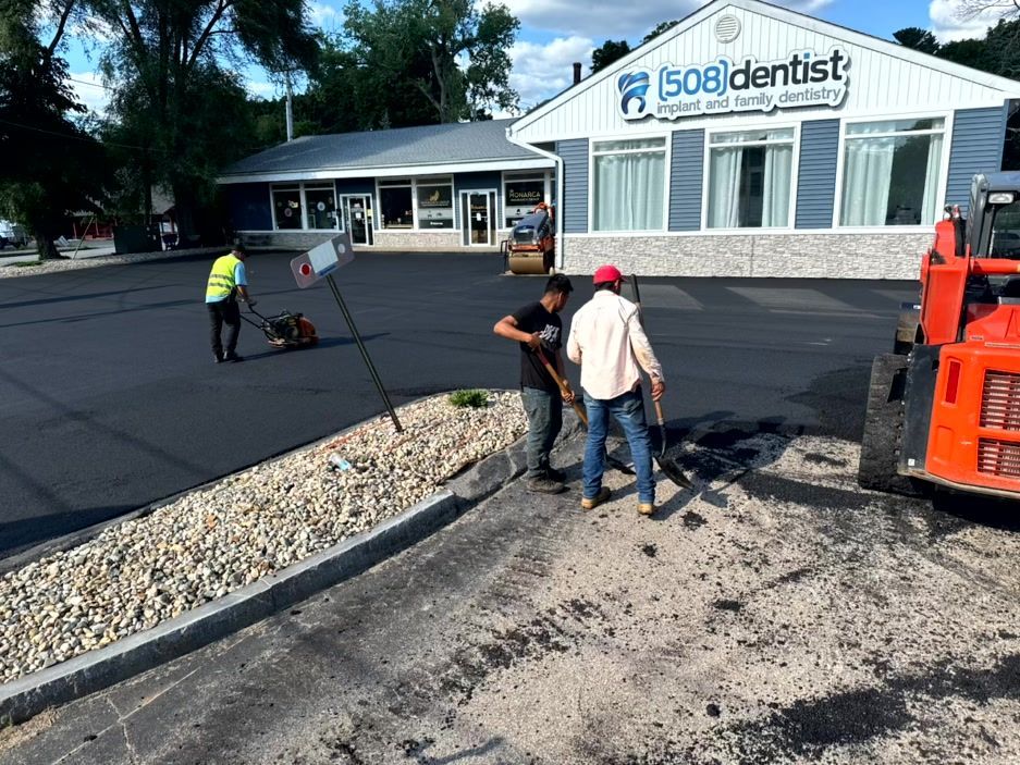 Asphalt paving work at a dentist's office. Workers spread asphalt, compact with a vibratory plate, and use tools.