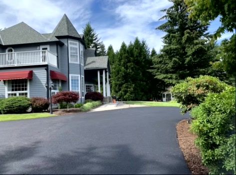 Gray house with turret, balcony, and red awning, set among trees and lawn, black paved driveway.
