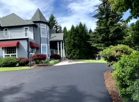 Gray Victorian house with a black driveway, red awning, and surrounding trees under a blue sky.