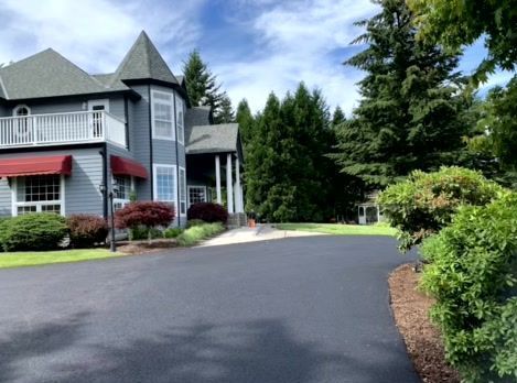 Exterior view of a gray Victorian house with a black driveway and lush landscaping under a partly cloudy sky.
