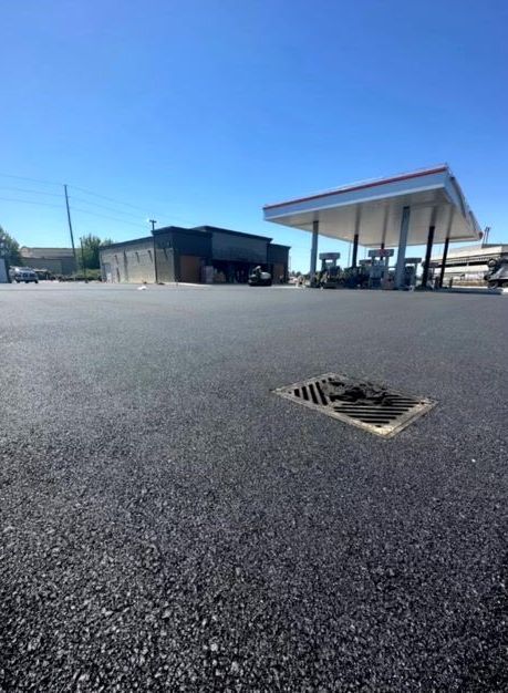 Gas station lot with new asphalt, building, and fuel pumps under a canopy on a sunny day.