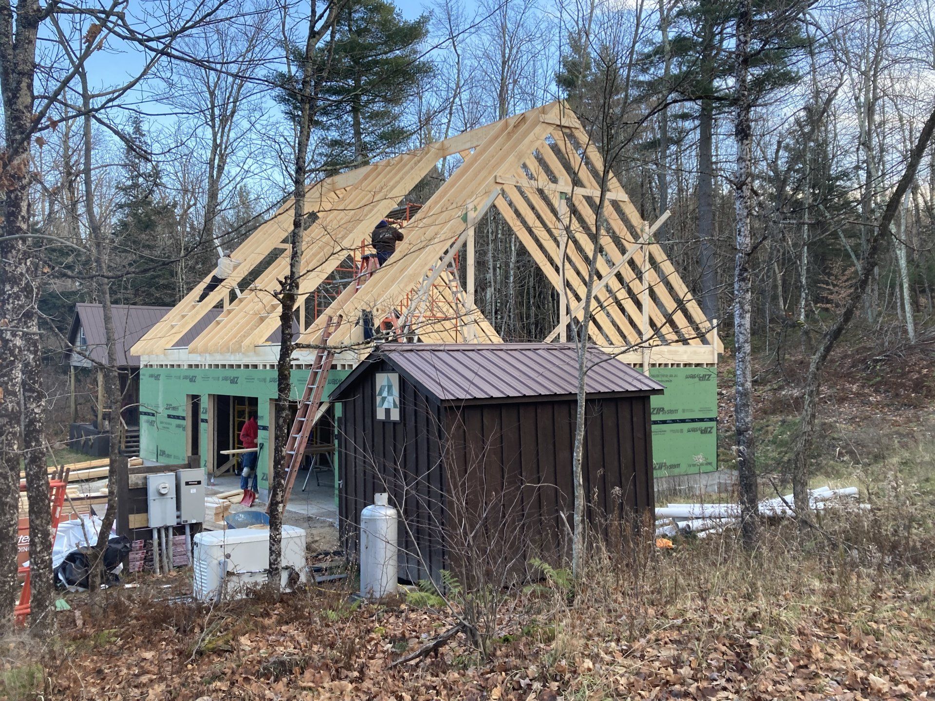 A house is being built in the woods with a shed in the foreground.
