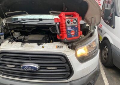 A white ford van with the hood open and a red jump starter attached to the hood.