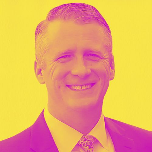 A man in a suit and tie is smiling on a yellow background.