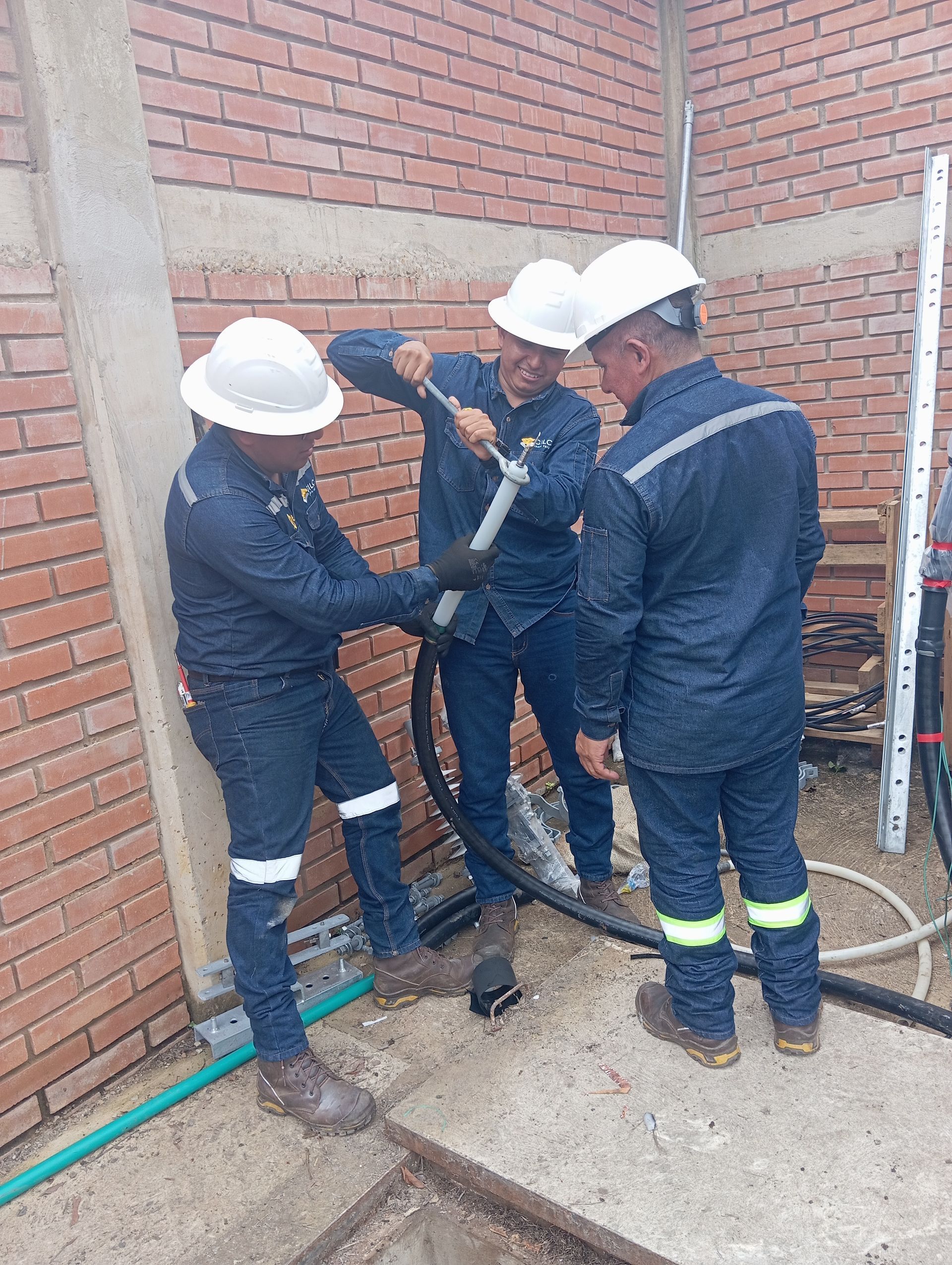 Tres trabajadores con cascos y monos de trabajo instalan conductos contra una pared de ladrillos en una obra en construcción.