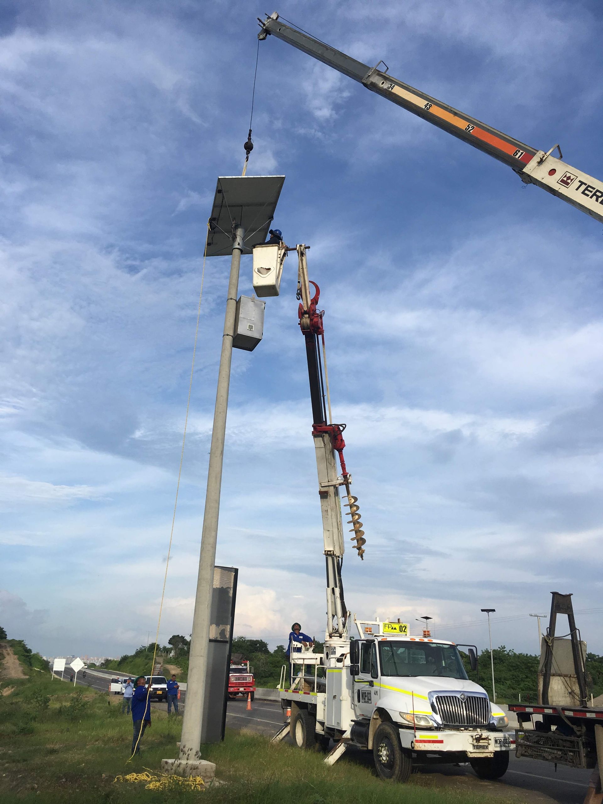 Una grúa eleva un panel solar situado en lo alto de un poste de servicios públicos, mientras que un camión con cesta elevadora facilita el acceso a los trabajadores que se encuentran junto a una carretera.