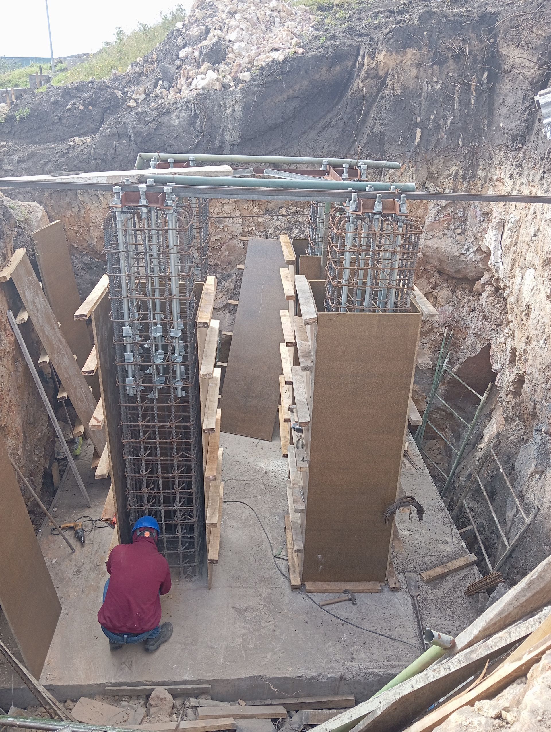 Un obrero de la construcción con una camisa roja se arrodilla junto a columnas de varillas de refuerzo y encofrados de madera dentro de una zanja de tierra.