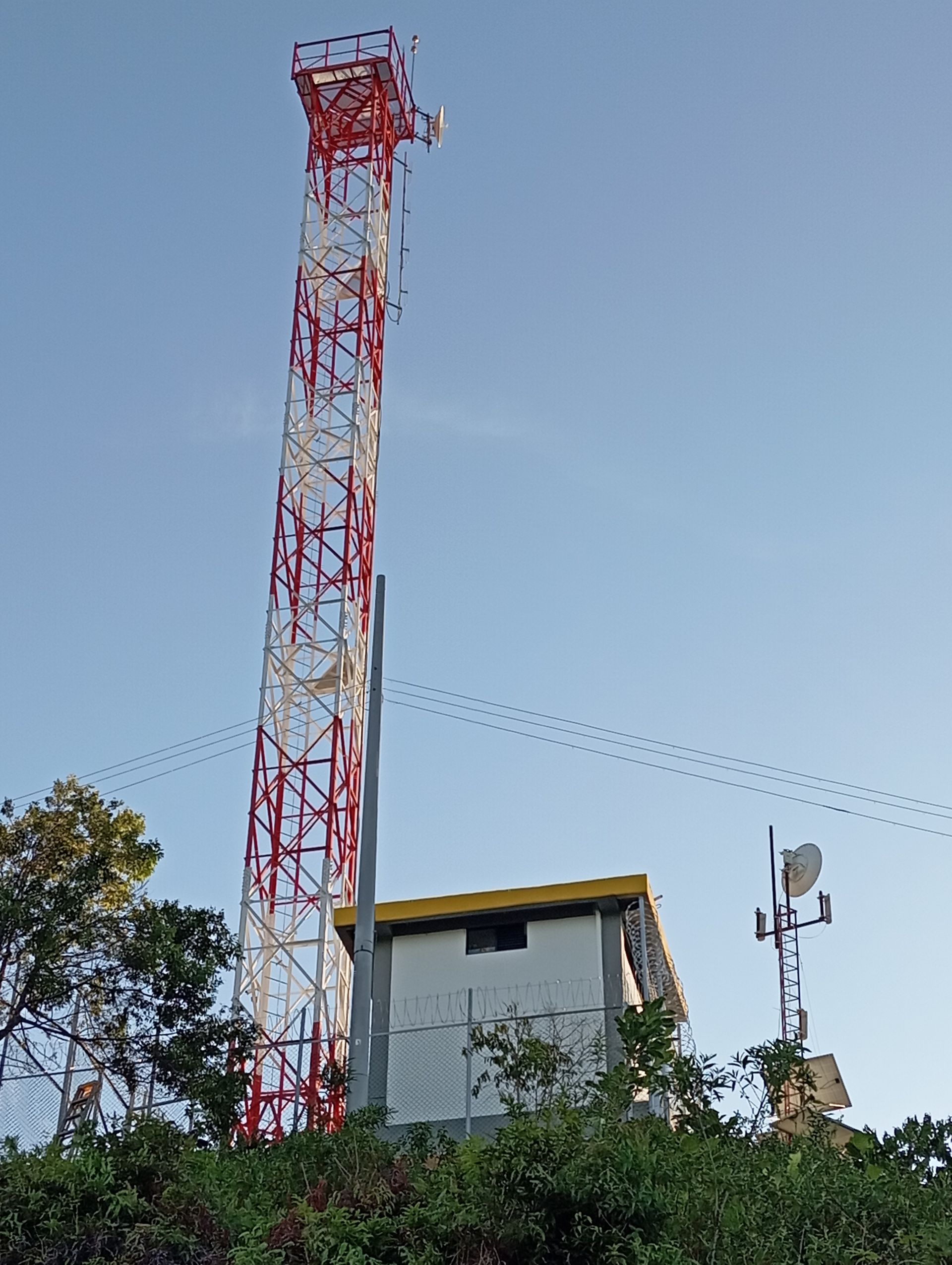 Una alta torre de comunicaciones metálica de color rojo y blanco se alza junto a un pequeño cobertizo para equipos en una colina bajo un cielo azul despejado.
