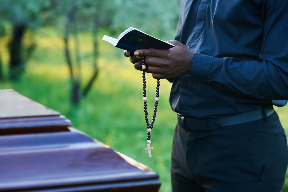 A man is holding a rosary and a bible in front of a coffin.