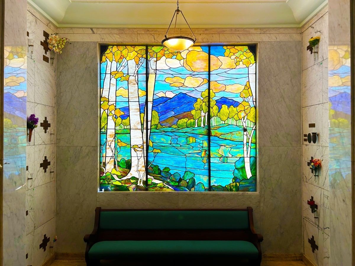 Interior of a mausoleum with a stained glass window depicting a landscape scene, a bench, and marble walls.
