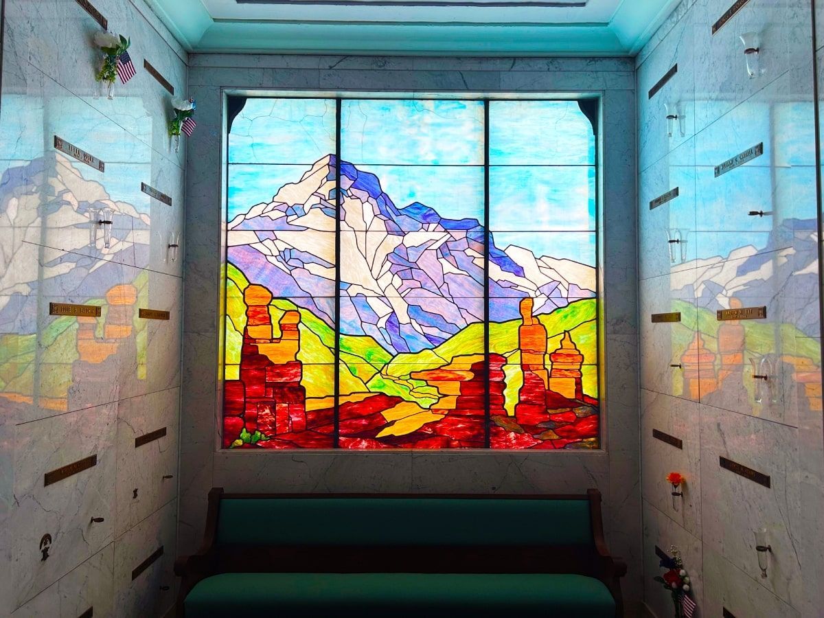 Stained glass window depicting a mountain scene inside a room with crypts and a teal bench.