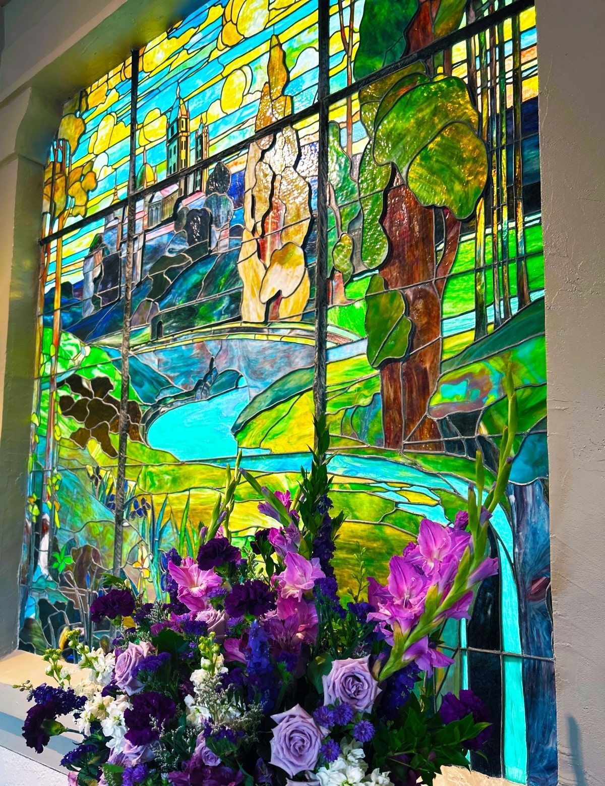 Stained glass window depicting a vibrant landscape; purple flowers in front.