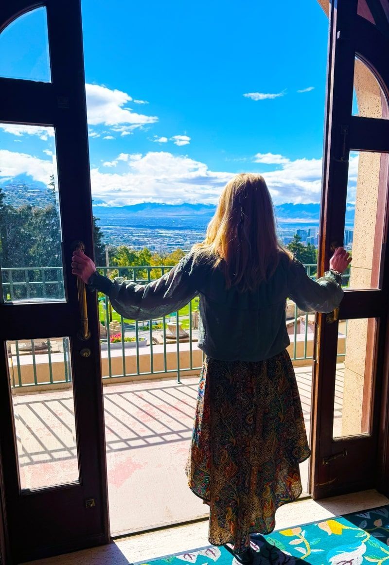 Woman looks out from arched doorway onto city and mountain view. She holds open doors.