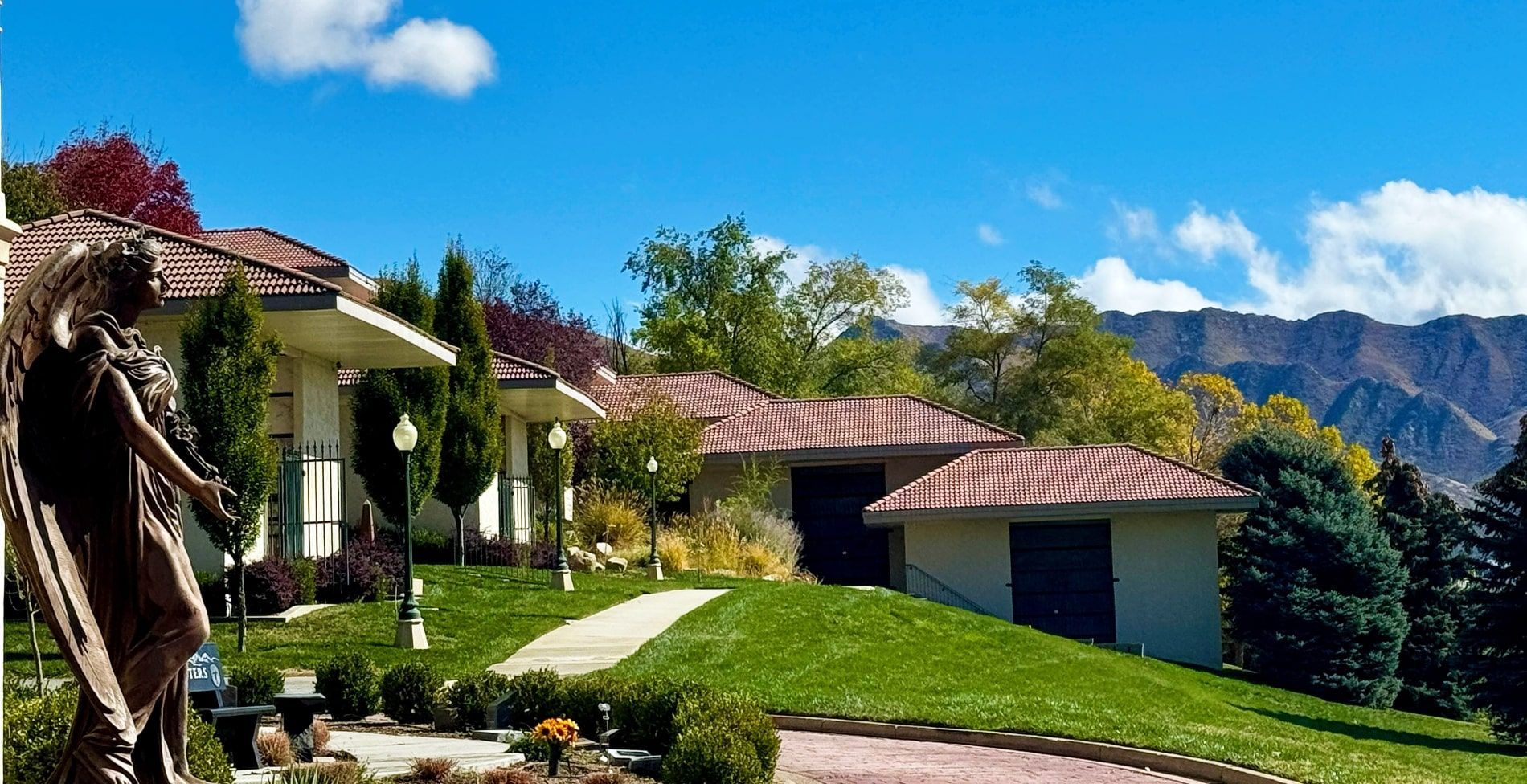 Bronze angel statue in front of a hillside building with red tile roof, lawn, and mountains.