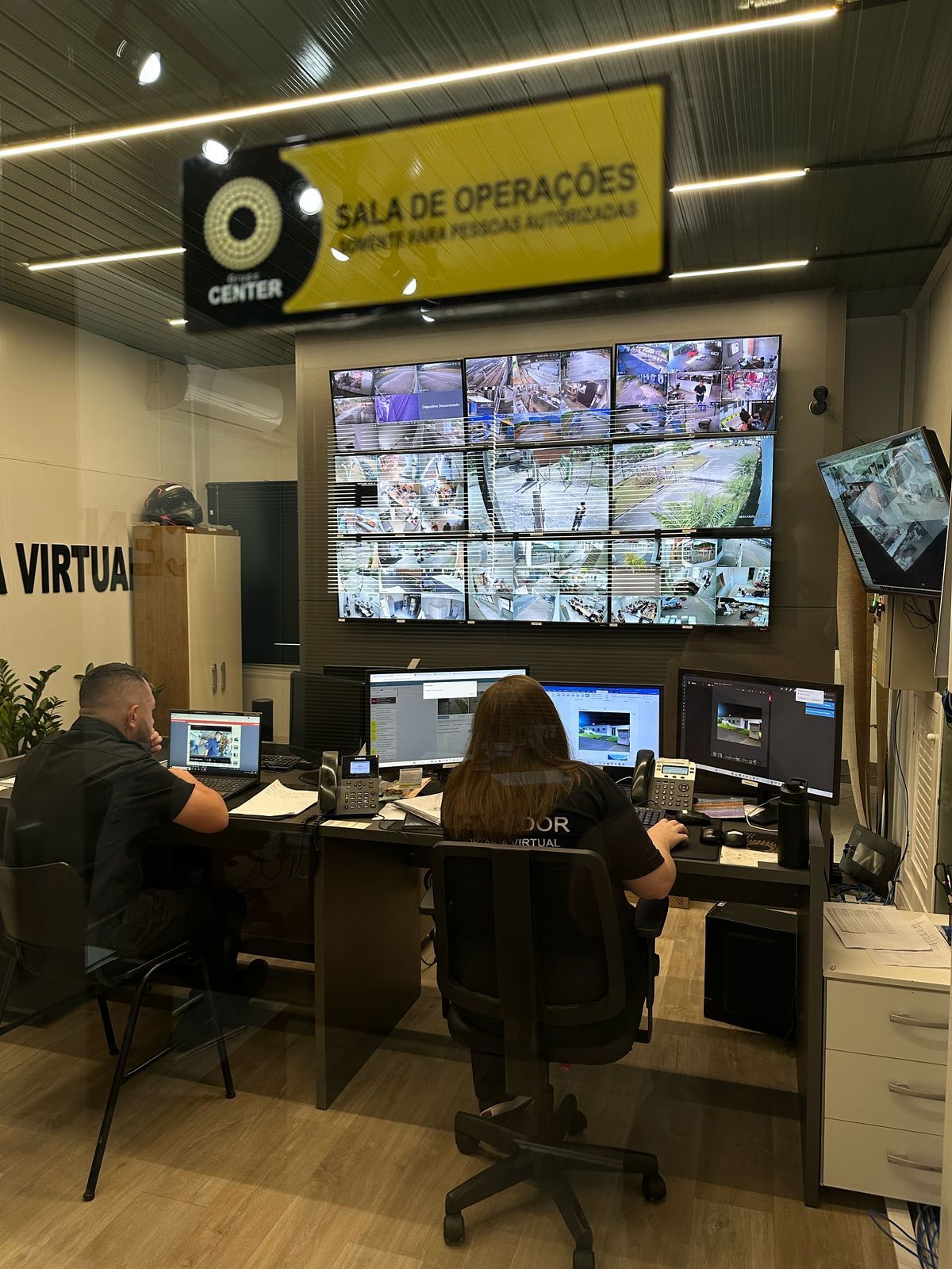 Duas pessoas em uma sala de operações de segurança monitorando várias telas. Uma placa no alto diz