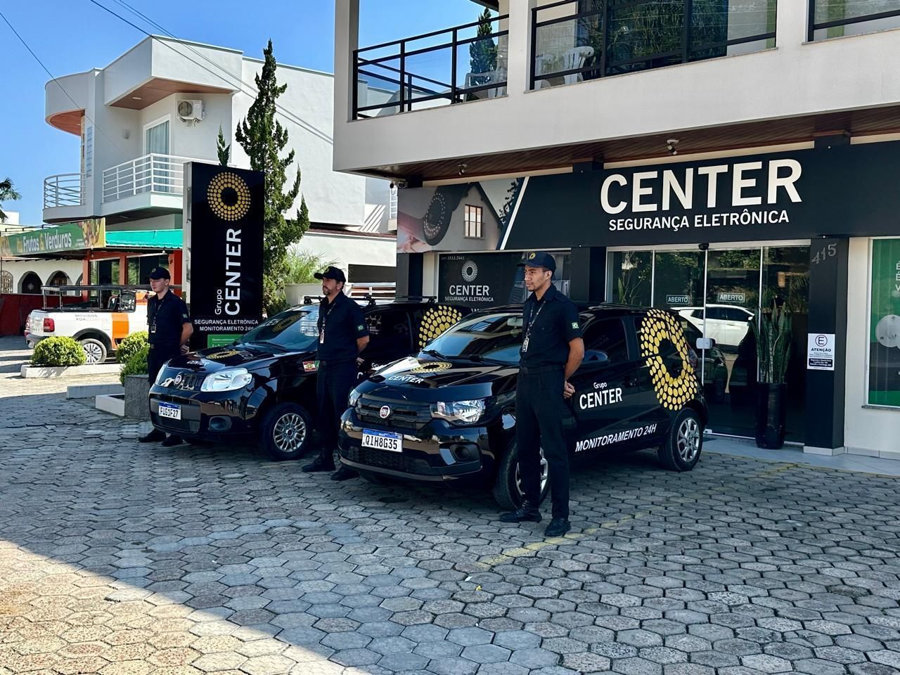 Três seguranças uniformizados estão ao lado de carros pretos com logotipos