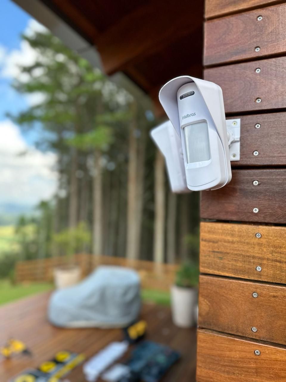 Um sensor de movimento branco com capa protetora está montado em uma parede de madeira. O fundo mostra um deck, árvores e ferramentas.