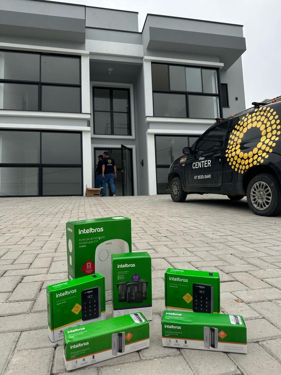 Exterior de uma casa com uma van preta próxima e várias caixas de produtos com etiquetas verdes e brancas na frente. Duas pessoas paradas na porta.