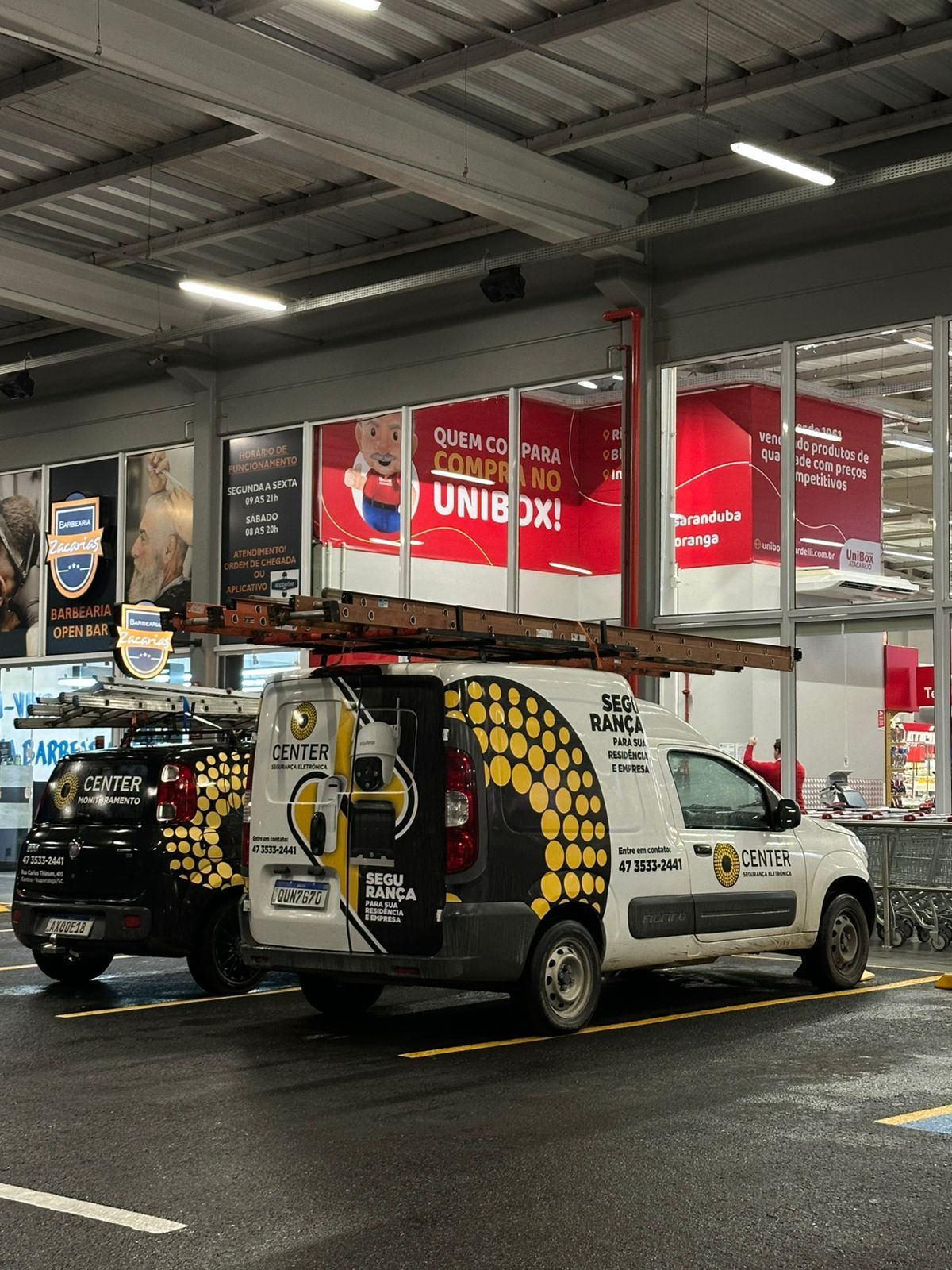 Duas vans de trabalho estacionadas em frente a uma loja. A van branca tem uma escada no topo e um padrão amarelo e preto.