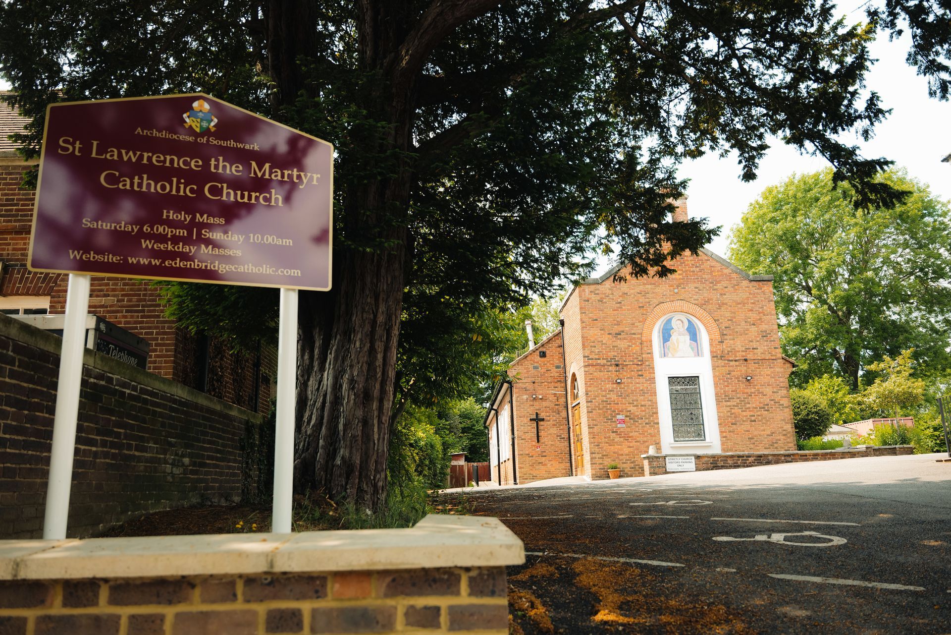 Sign for St. Lawrence the Martyr Catholic Church next to a brick building with a tree in front.