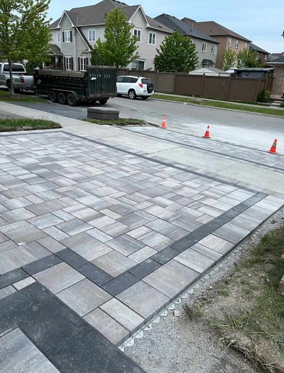 A newly installed paver stone driveway with a dark gray border pattern, viewed from the sidewalk in a suburban neighborhood.