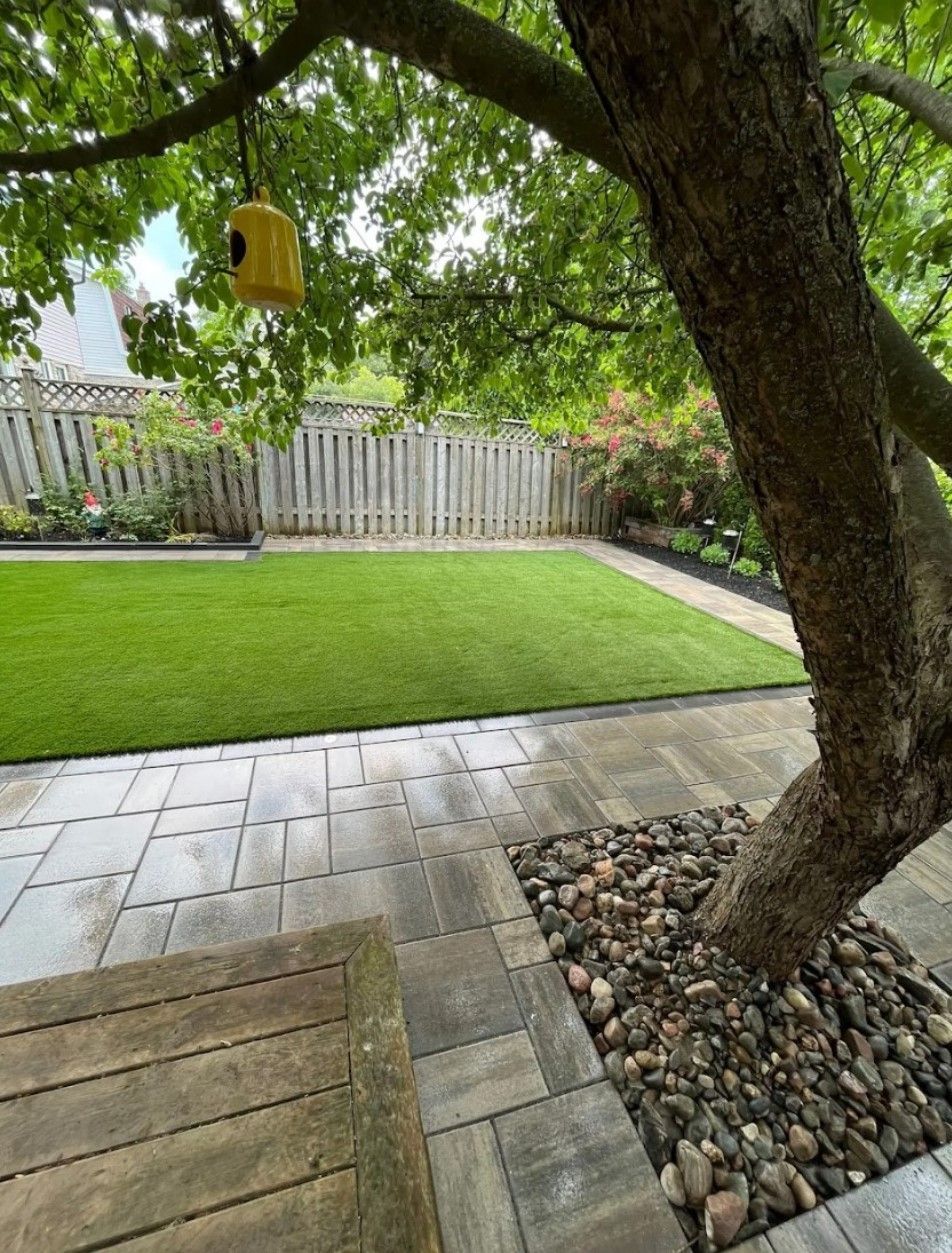 A backyard featuring a stone patio, a patch of green grass, a wooden fence, and a tree with a birdhouse hanging from it.