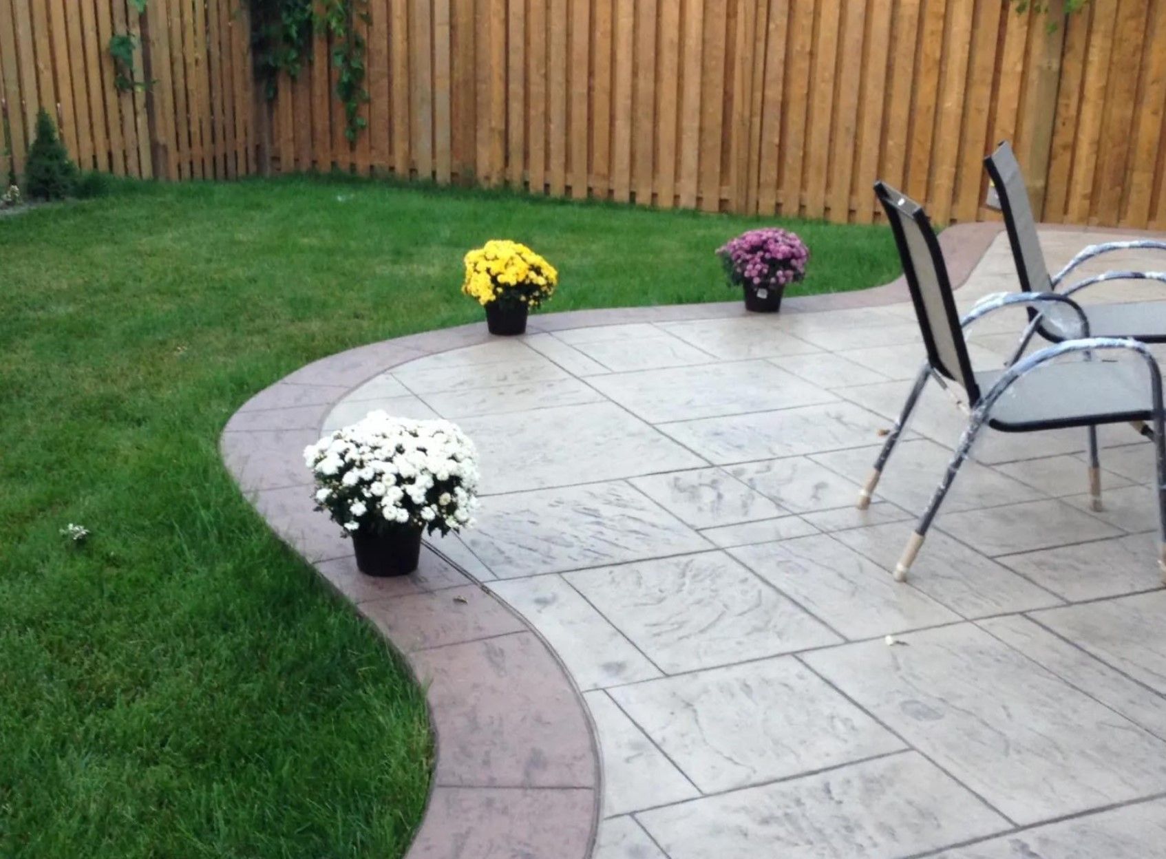 A stamped concrete patio with curved edges, three pots of flowers, and two chairs in a backyard with a wooden fence.