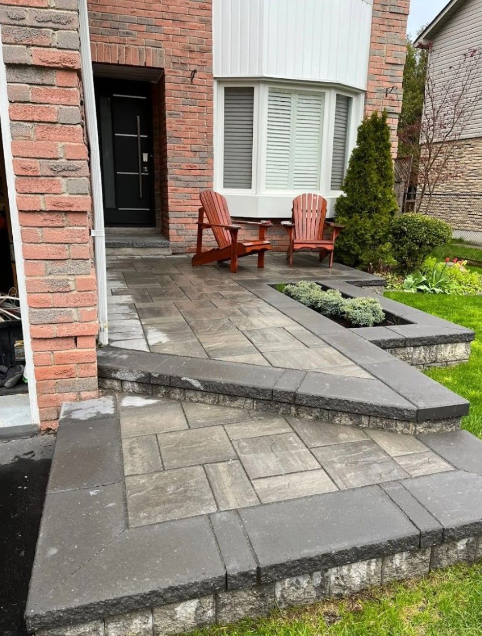 A patio with gray stone pavers and a raised garden bed leads to a brick house entrance with two wooden Adirondack chairs.