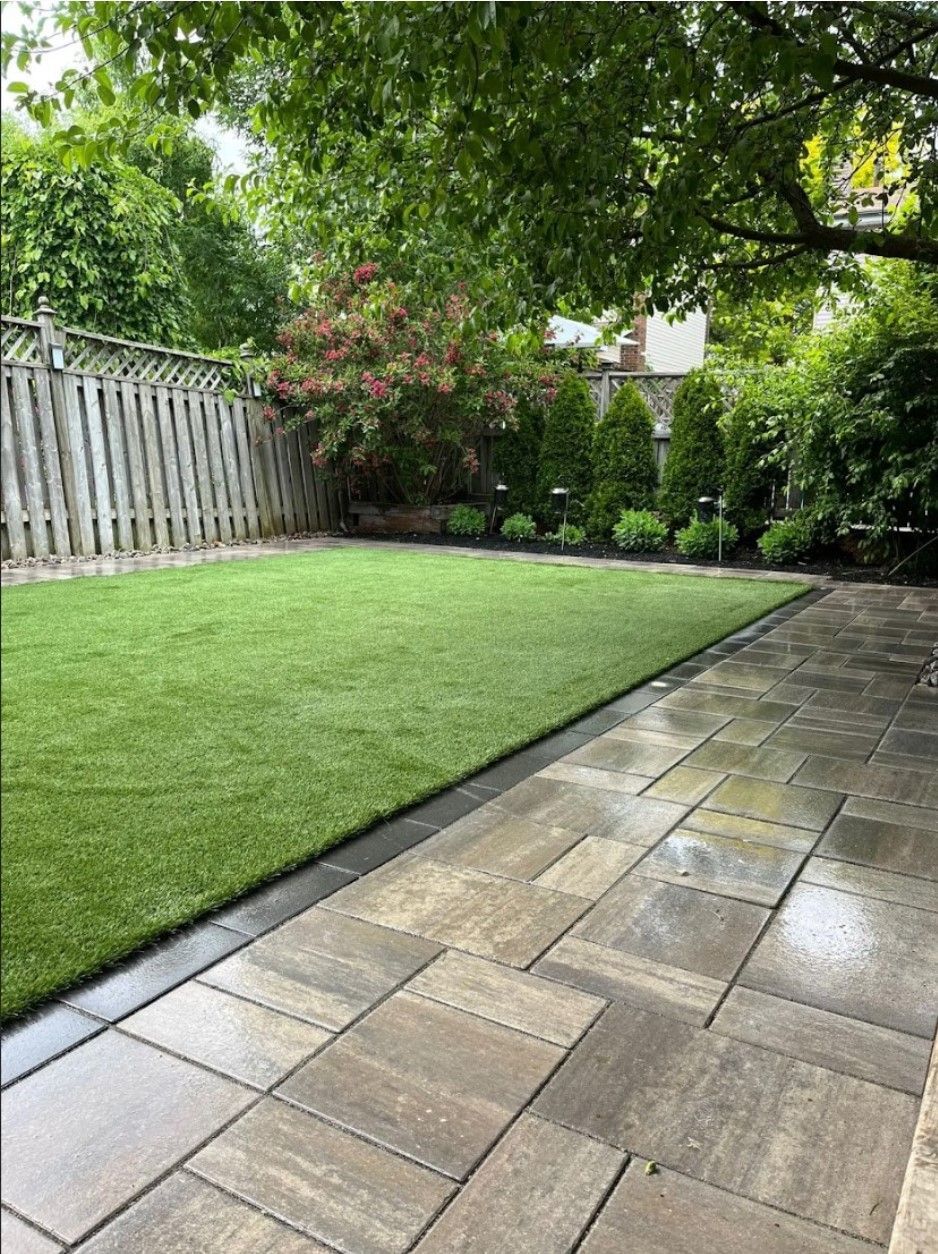 A paved patio borders a neat green lawn next to a wooden fence and various shrubs and trees under a leafy canopy.